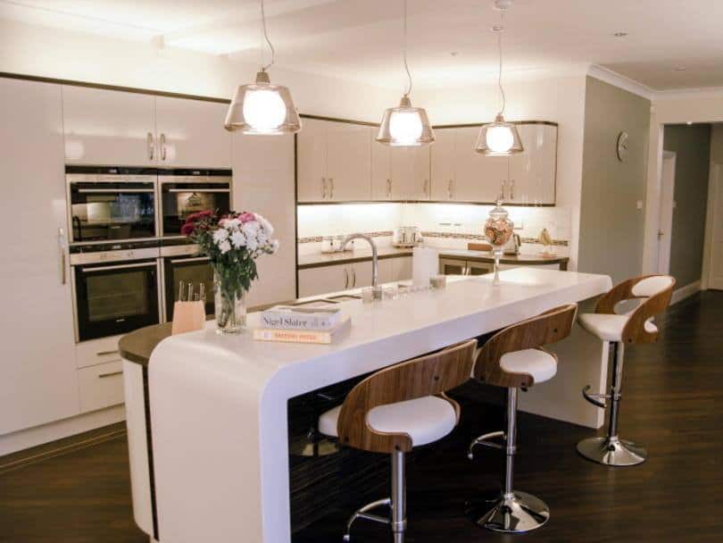 seating area for three at a bar with thick cream corian worksurface above tall units with ovens in to left hand side and three pendant lights over the island, Beige And Ebony,