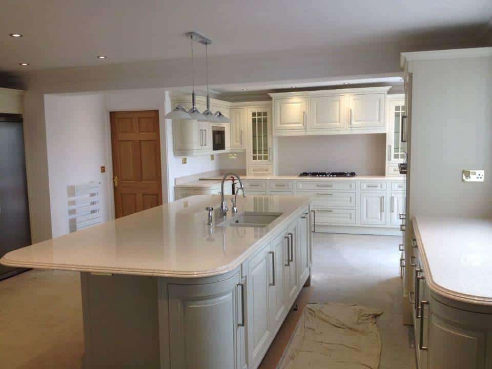 An Astor kitchen with an island and sink , having curved cabinets on each side light coloured doors and light worksurface reflect the light back into the room, a traditional mantle is shown to the rear with a hob underneath