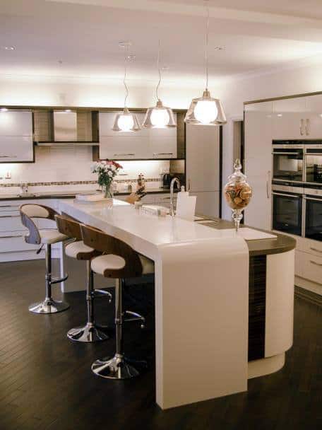 seating area for three at a bar with thick cream corian worksurface above tall units with ovens in to left hand side and three pendant lights over the island