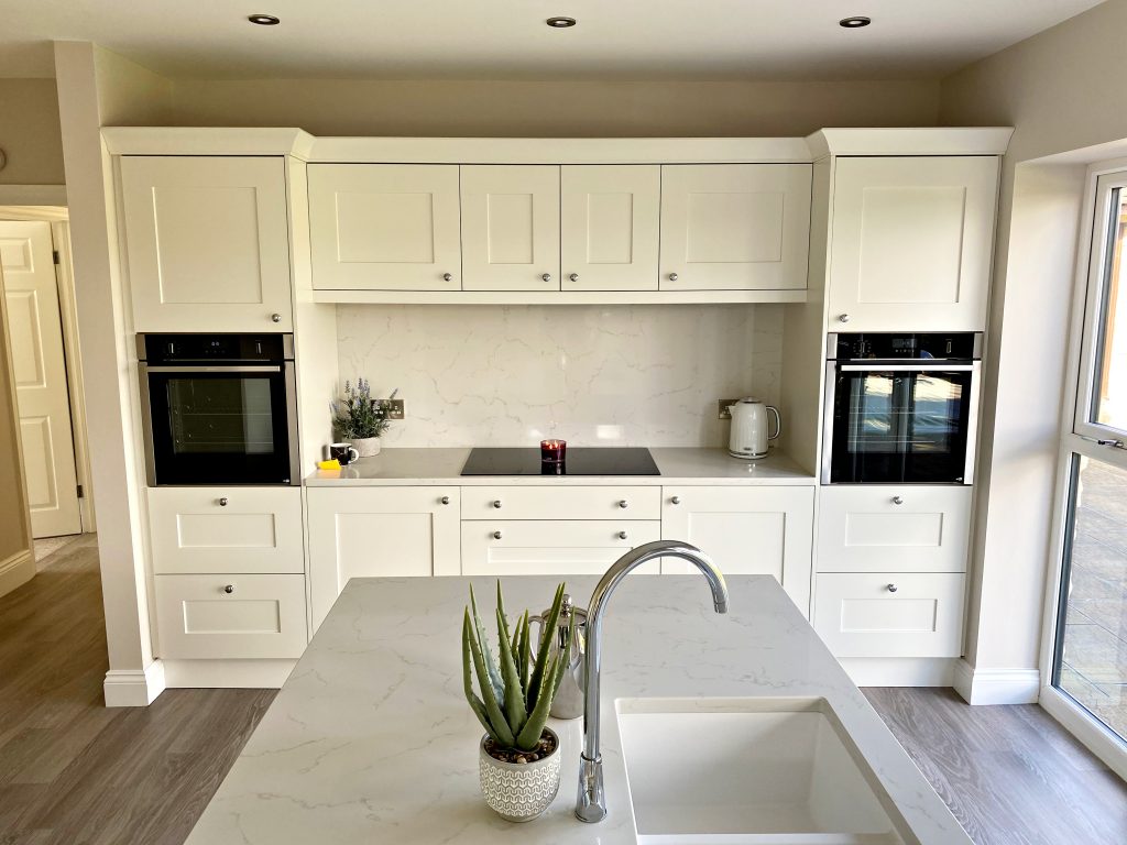 fitted kitchen design in Teesside with a oak texture painted in off cream , an oven tower is either side of a small run of base cabinets with a hob in the center , an island with a sink in is to the foreground