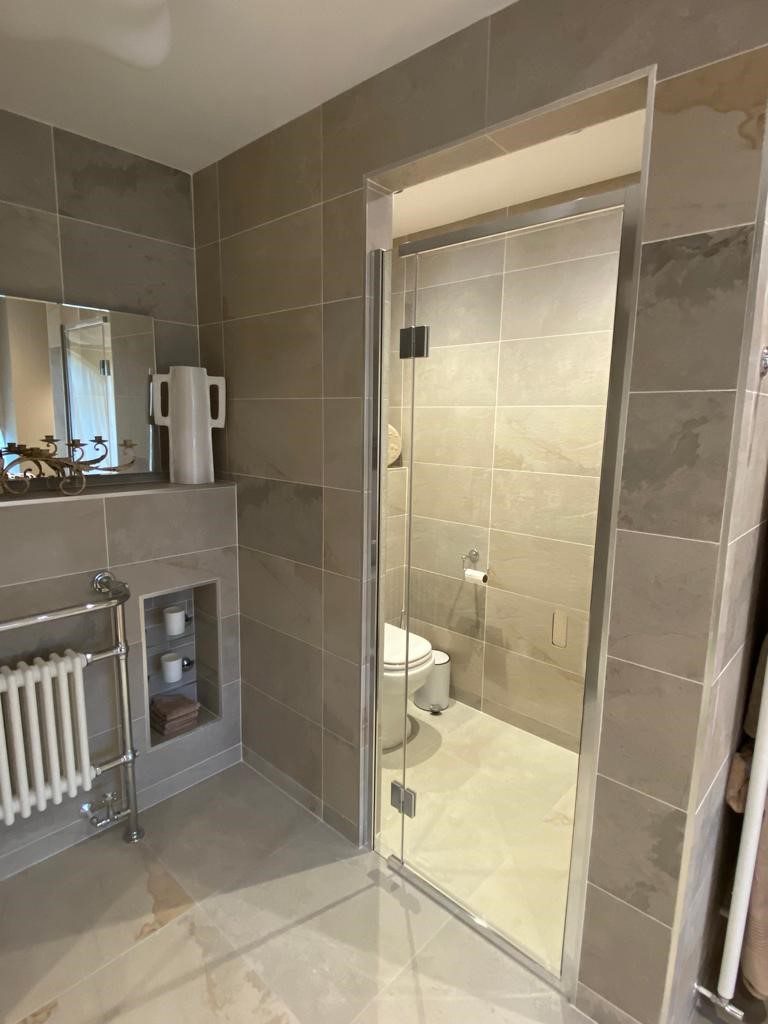 Fully tiled bathroom with glass door leading to floor mounted white toilet, to the left a traditional white radiator below a mirror